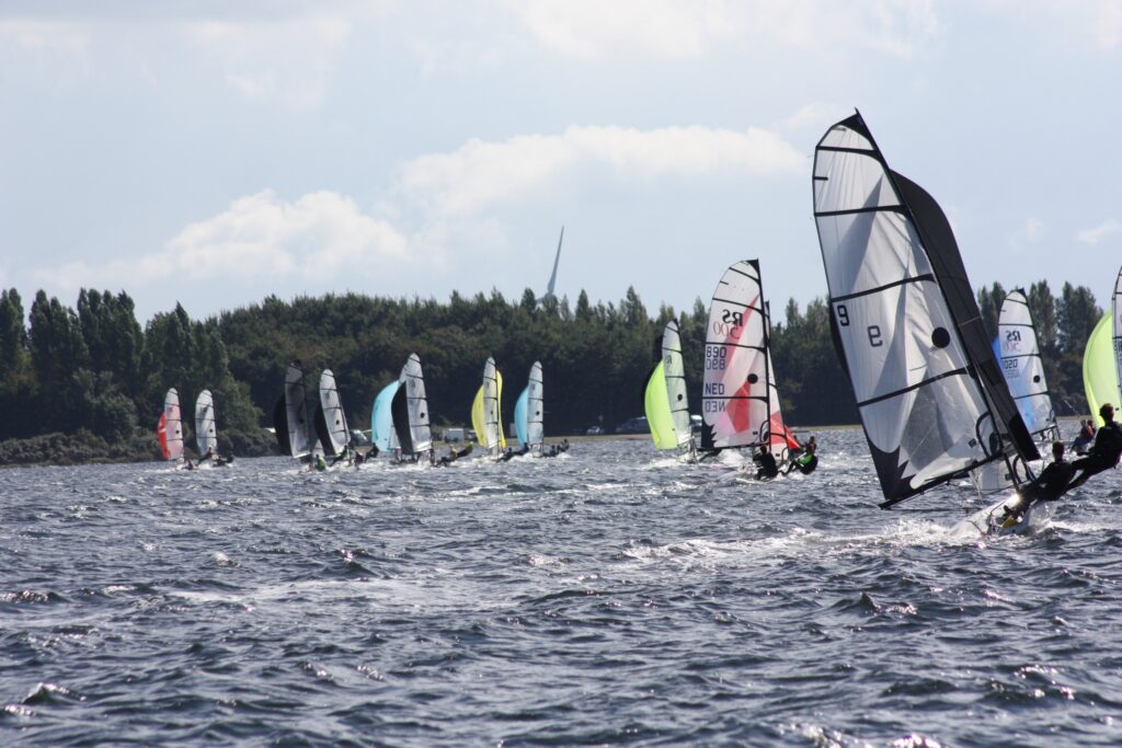 Sailing dinghies race in choppy water with colorful sails near a tree-lined shore.