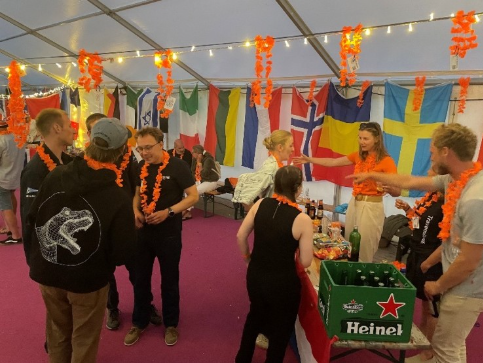 People mingle at a tented sailing event with flags from multiple countries, orange garlands, and a Heineken bar setup.