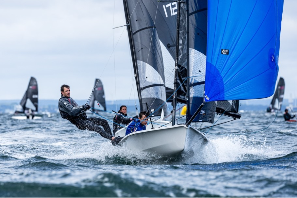 Sailing dinghy with crew leaning out as sails catch wind near bow, blue spinnaker on right.